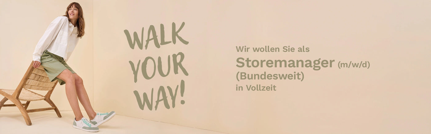 Eine lächelnde Frau mit Brille steht in einem hellen Schuhgeschäft mit Regalen voller Schuhe im Hintergrund.