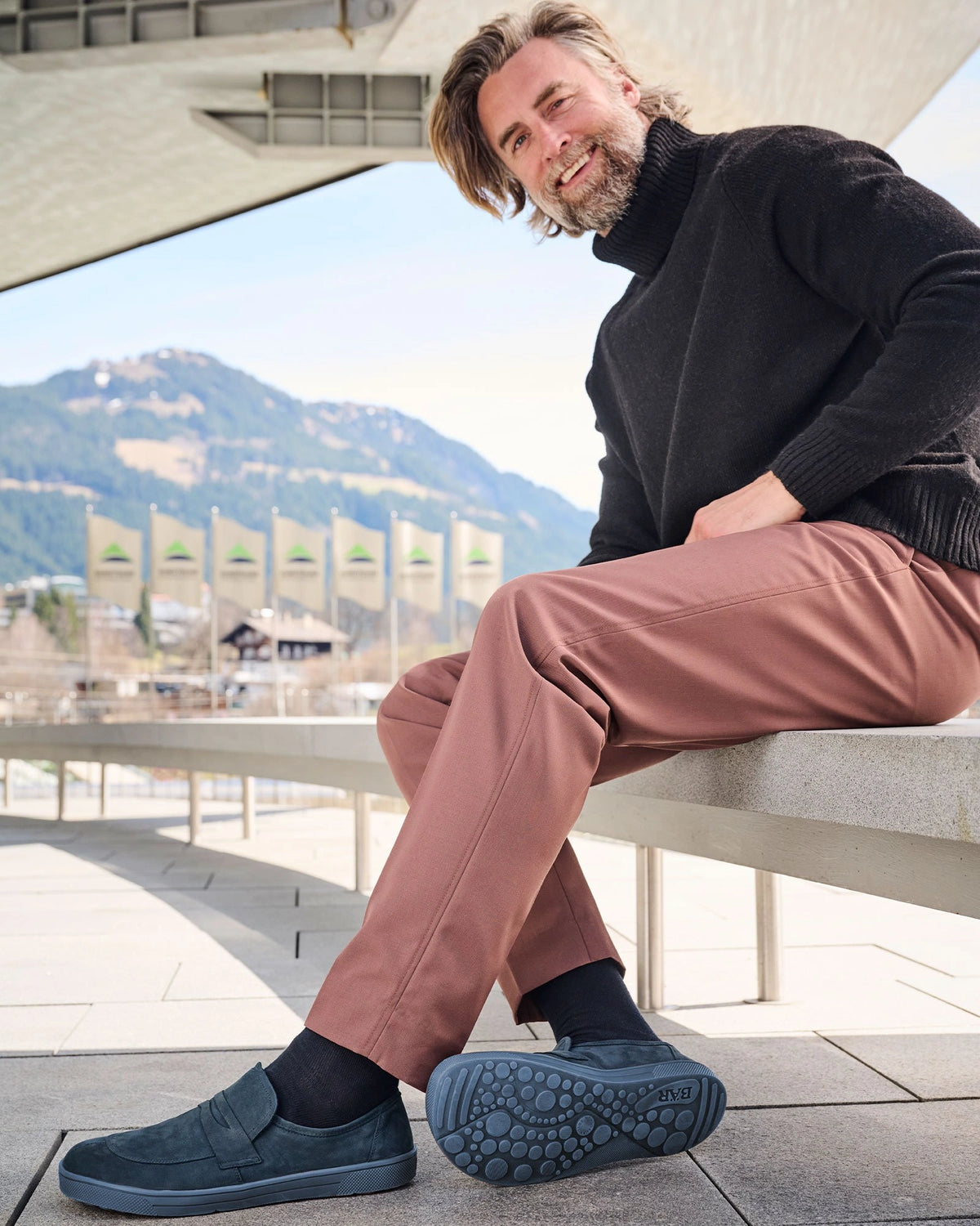Lächelnder Mann sitzt auf einer Terrasse, trägt einen schwarzen Pullover, braune Hose und blaue Slipper. Im Hintergrund sind Berge und Fahnen sichtbar.