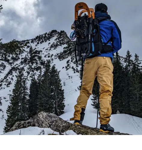 Mann in blauer Jacke und gelber Hose mit Schneeschuhen im Rucksack vor verschneiter Bergkulisse