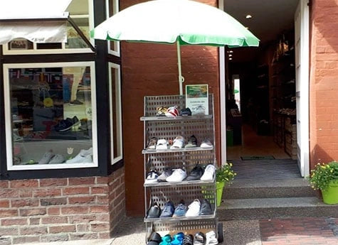 Ein Schuhständer vor einem Geschäftseingang, geschützt von einem grünen Sonnenschirm, mit verschiedenen Sportschuhen ausgestellt.