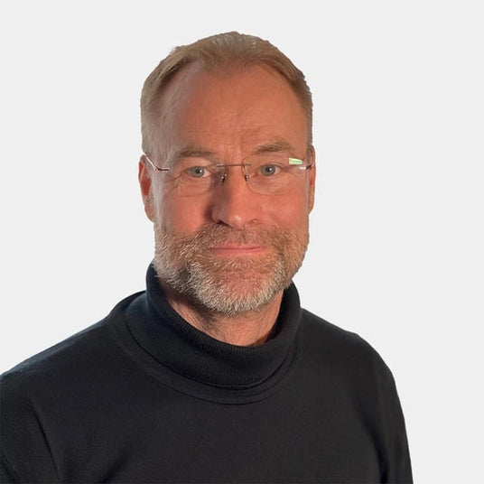 Ein lächelnder Mann mittleren Alters mit Brille und Bart, trägt einen schwarzen Rollkragenpullover vor einem neutralen Hintergrund