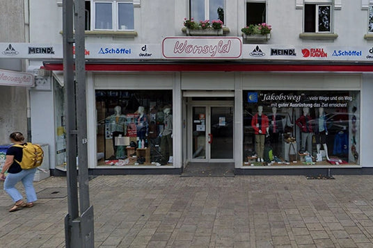 Eingangsbereich eines Outdoor- und Bekleidungsgeschäfts mit Schaufenster und Markenlogos wie Meindl und Schöffel. Eine Person mit gelbem Rucksack geht vorbei.
